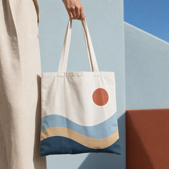 Person holding a cream canvas tote bag printed with a sun and wave design in terracotta, blue and beige.